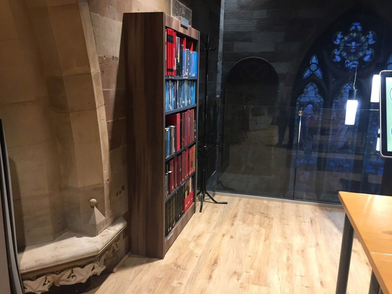 Dark wood shelving unit with red and blue books in a study area beside a stone wall at Lichfield Local Studies