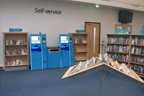 Two blue self-service kiosks beside a face-out book display and browsing shelves at Patchway Library