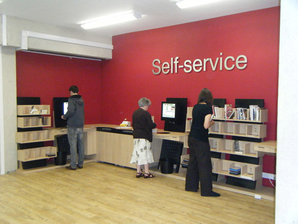 Three patrons using self-service kiosks at a dedicated self-service area with wooden shelving at Oxfordshire Central Library