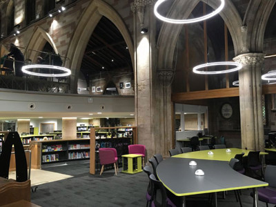 Circular pendant lights illuminate a modern study area with green and grey tables and pink lounge chairs at Lichfield Library