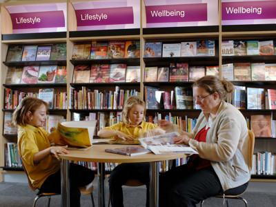 Two children in yellow shirts and an adult seated at a round table, engaging with books in a wellbeing browsing area at Far Cotton Library
