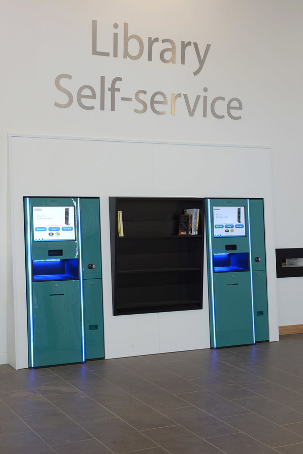 Two self-service kiosks with illuminated screens and a black shelving unit for returns at Southwater Library