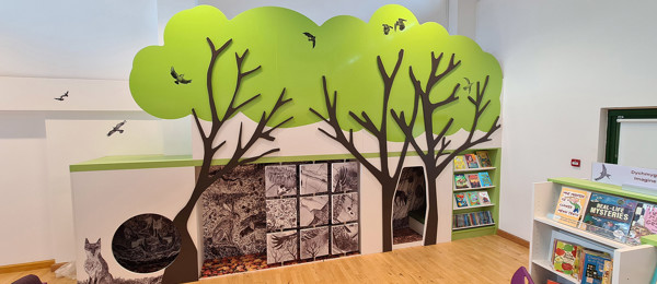 Colourful tree-themed wall graphics and face-out book displays in a children's reading area at Llanrwst Library