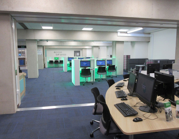 Curved wooden desks with computers and black office chairs in a self-service area at Oxfordshire County Library