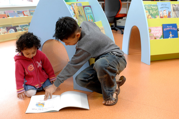 A boy pointing at a picture book while sitting beside a girl in a children's reading area with colourful book displays at High Street Library Bolton