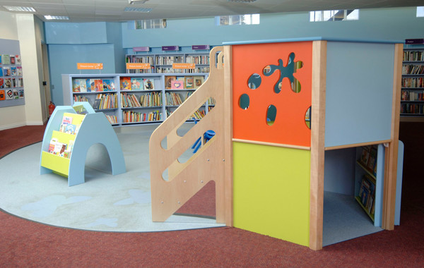 Colourful play structure with slides and cut-out shapes beside a book display unit in the children's reading area at Sandsend Library