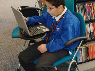 A student using a laptop on a portable desk in a study area with browsing shelves at Djanogly Academy