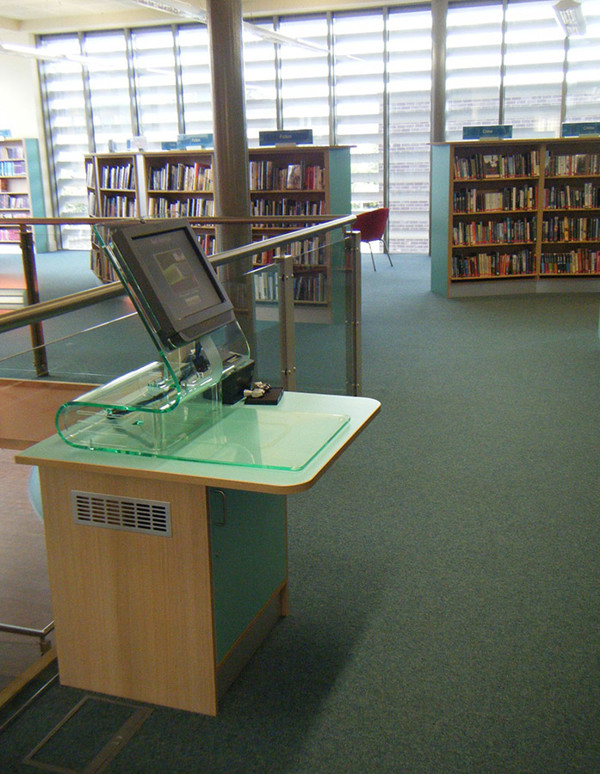 Self-service kiosk with a clear housing unit beside browsing shelves at Failsworth Library