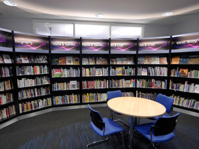 Curved black shelving units filled with books and a round wooden table with blue chairs in a browsing area at Thame Library