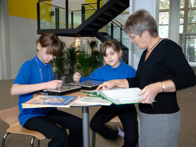 Two students engaged with books and a tablet at a collaborative work table in a study area at Djanogly City Academy