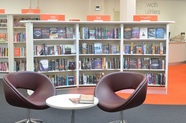 Two burgundy lounge chairs and a white side table beside browsing shelves filled with books at Llandudno Library