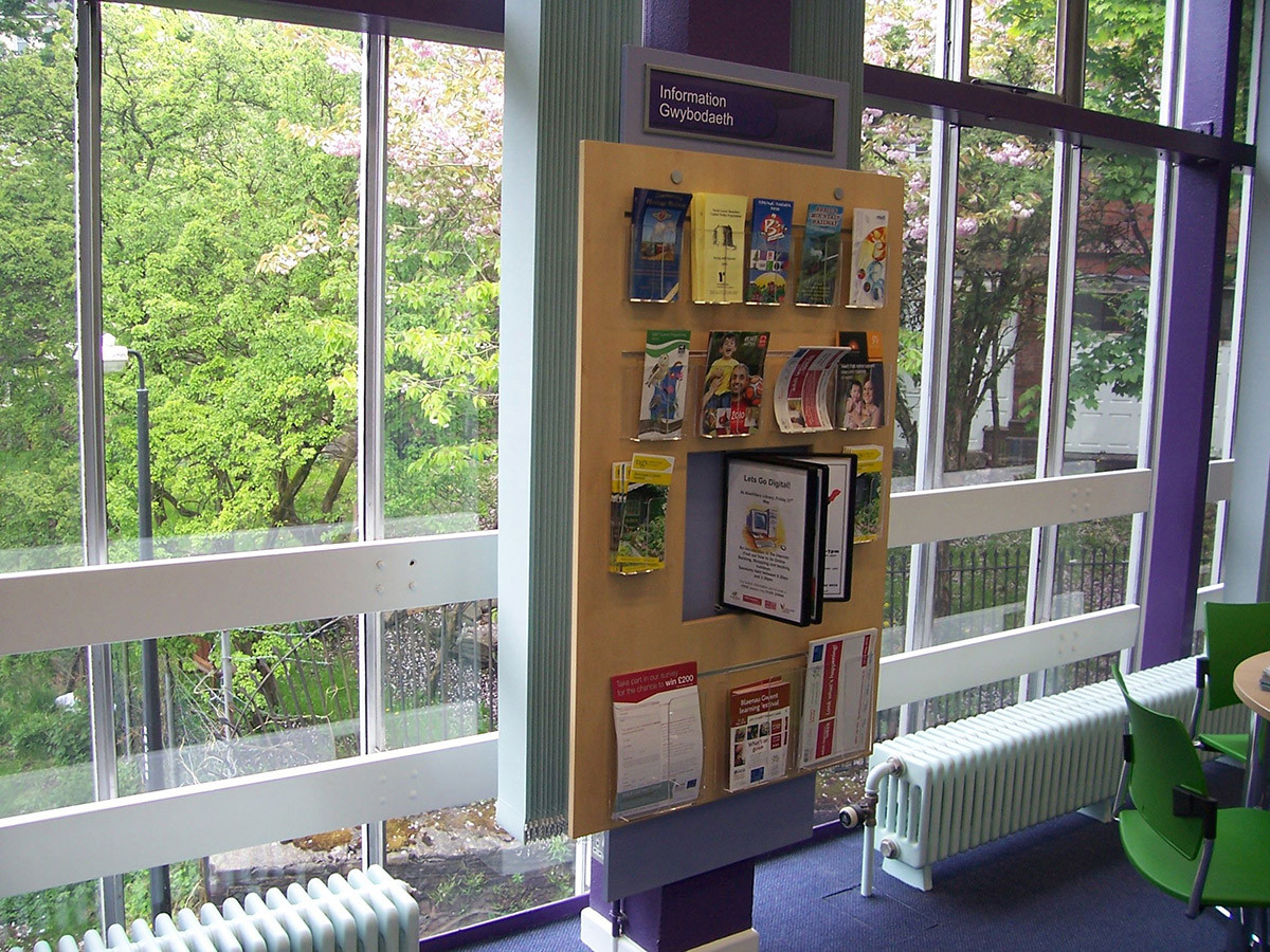 Wooden leaflet display unit with various brochures and information guides beside large windows at Abertillery Library