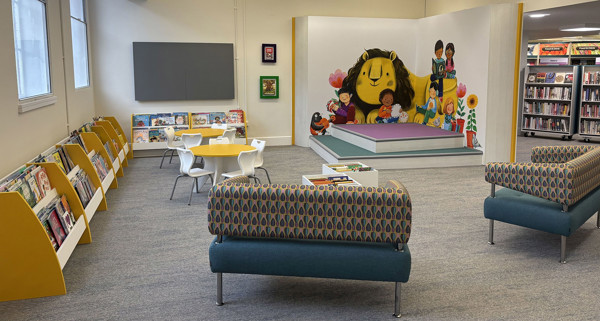 Curved blue sofas and a yellow table in a children's reading area with a vibrant mural at Sale Library