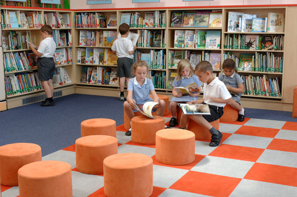 Orange cylindrical stools arranged on a checkered carpet in a children's reading area, with children browsing books at Wentworth Primary School