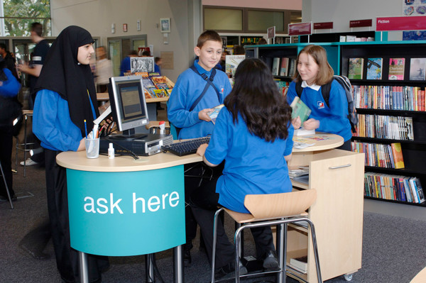 Students engaging with staff at a modern help desk featuring a circular counter and browsing shelves at Djanogly City Academy Library