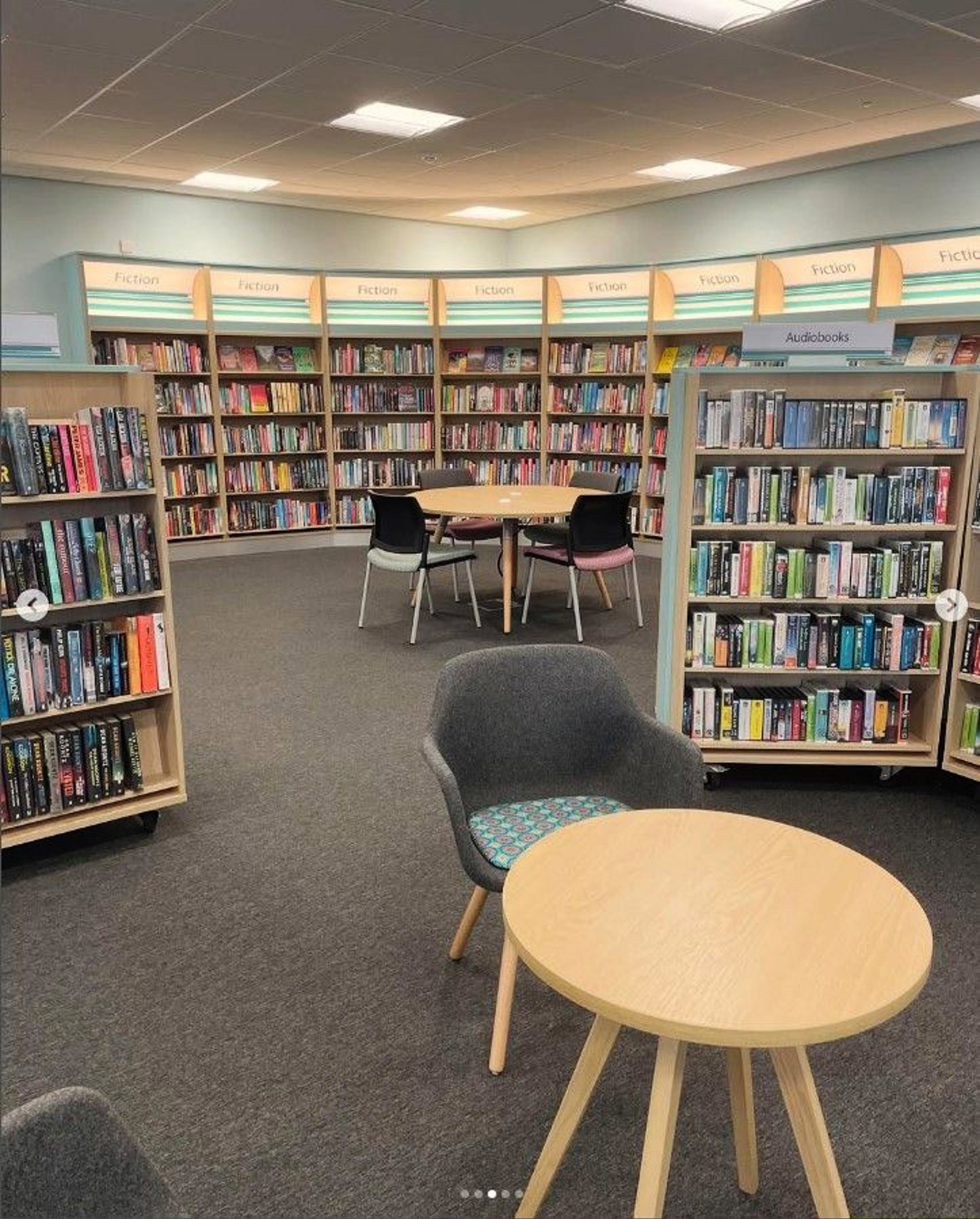 Grey upholstered chairs and light wood tables in a public library browsing area with fiction and audiobook shelves at Birtley Library