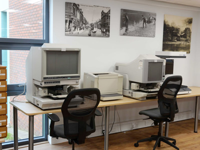 Two desktop computers with ergonomic chairs beside archival storage drawers and historical photographs at Gateshead Archive