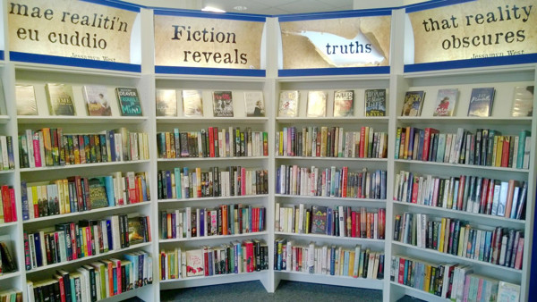 Curved shelving displaying a diverse collection of books with thematic signage at Ystrad Mynach Library