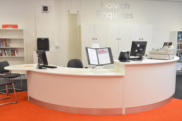 Curved white help desk with computer stations and an information display on an orange carpet at Llandudno Library