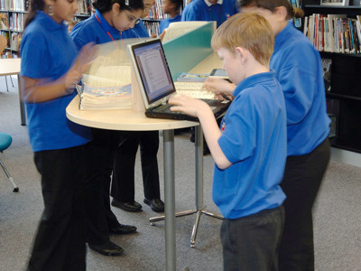 Students in blue shirts collaborating at a study table with laptops and resources in a library browsing area at Djanogly Academy