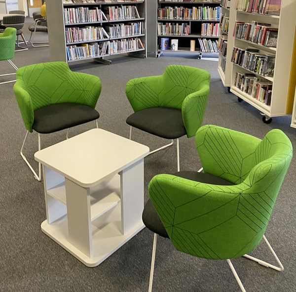 Green upholstered lounge chairs surrounding a white side table in a reading area at Holt Park Library