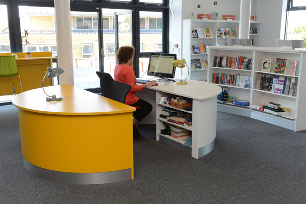 Curved yellow help desk with a seated staff member using a computer beside browsing shelves at King Edward's School Library