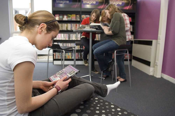 A young girl reading a book on a floor cushion while three peers engage in discussion at a study table at Morpeth Library