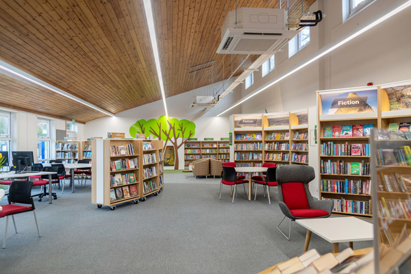 Red and grey lounge chairs beside browsing shelves and a tree mural in a public library browsing area at Chinnor Library