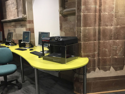 Bright yellow curved desk with a scanner and computer stations against a stone wall at Lichfield Library