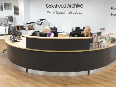 Curved reception desk with staff assisting visitors at Gateshead Archive, featuring a modern design and wooden flooring at Gateshead Archive