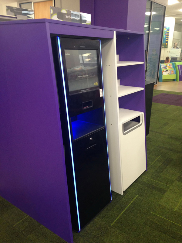 Self-service kiosk with a sleek black design and blue lighting beside a white drop bin at The Place Library & Theatre Telford