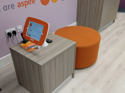Orange pouffe and a wooden side table with a digital information screen in a collaborative work area at Newcastle-under-Lyme Library