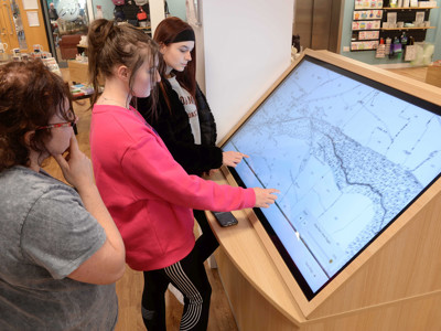 Three young adults interacting with a large touchscreen display featuring historical maps in the local studies area at Gateshead Archive