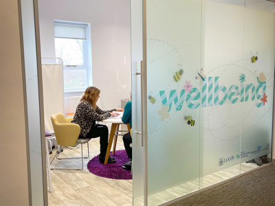 Glass partition with "wellbeing" graphic and two people seated at a study table in a quiet reading space at Leeds Wellbeing Pods