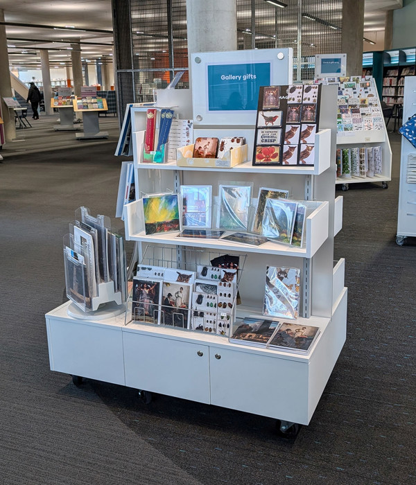 Mobile display unit featuring gallery gifts, showcasing cards and art prints in a public library browsing area at Oldham Library & Gallery Shop – gallery gifts