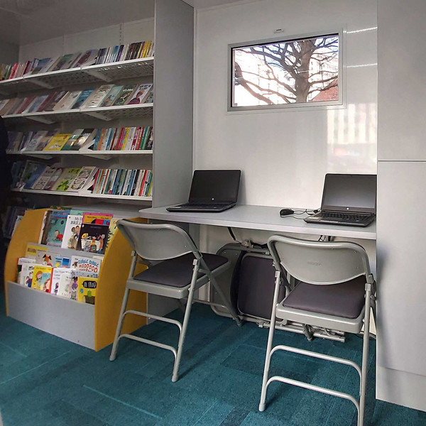 Two grey folding chairs and a study table with laptops in a public library browsing area at Leeds Mobile Community Hub