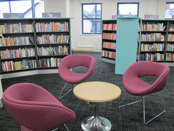 Pink lounge chairs and a round wooden table in a reading lounge beside browsing shelves at Caerphilly Library