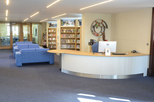 Curved reception desk with a computer and blue seating area beside browsing shelves at Kingham Hill School Library