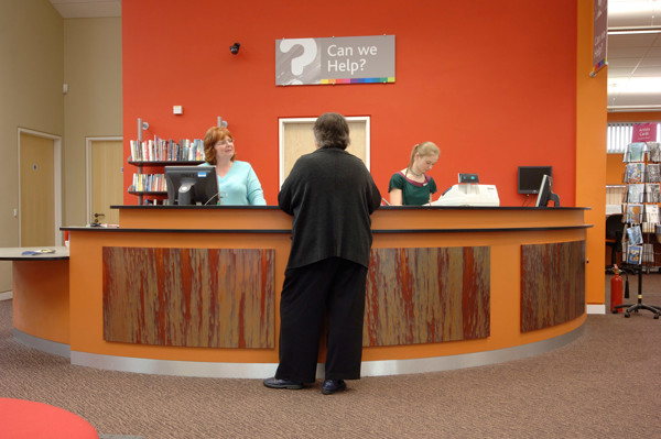 Curved help desk with a wooden finish and orange wall behind, featuring staff assisting a visitor at Longlevens Library