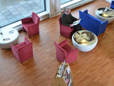 Red and blue upholstered lounge chairs with circular tables featuring artistic designs in a public library browsing area at Risca Palace Library