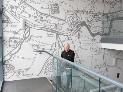 A man standing on a glass staircase beside a large historical map mural at Gateshead Archive