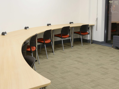 Curved wooden study tables with power access and black chairs featuring orange upholstery in a collaborative work area at Brackenhurst Campus Library