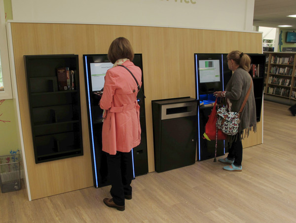 Two patrons using self-service kiosks with integrated return shelves and drop bins in a modern library space at Newark Library