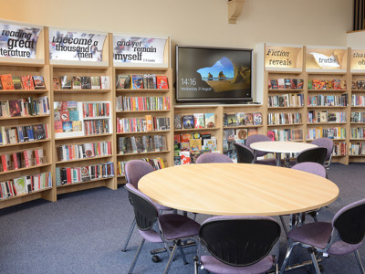 Circular tables surrounded by upholstered chairs beside browsing shelves and a screen at Kingham Hill School