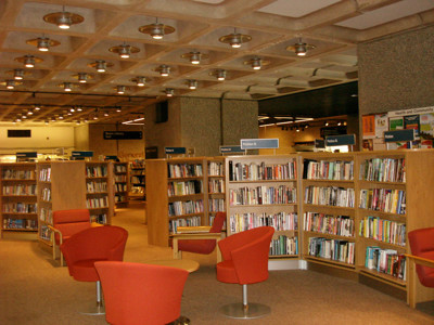 Orange tub chairs and circular tables beside wooden browser shelving in a public library browsing area at Barbican Library