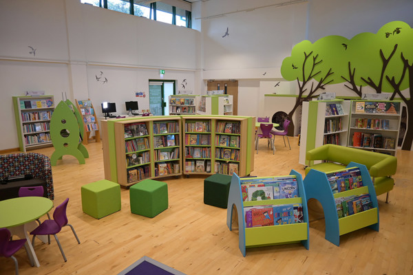 Bright green and blue book displays alongside circular seating and colourful stools in a children's reading area at Llanrwst Library