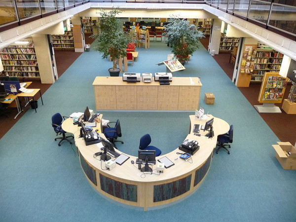 Curved help desk with multiple computer stations and blue chairs in an open-plan library space at Oxford Central Library
