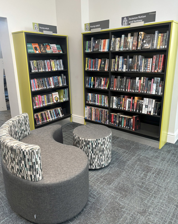 Curved grey upholstered seating and circular ottoman beside black shelving filled with young adult titles at Royton Library