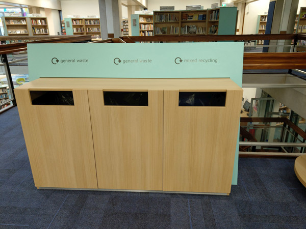 Wooden recycling and waste bins with clear signage in a public library browsing area at County Library, Oxfordshire