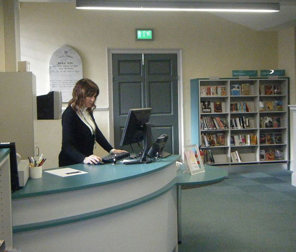 Curved help desk with a computer and a staff member assisting patrons beside browsing shelves at St Aubyn Library